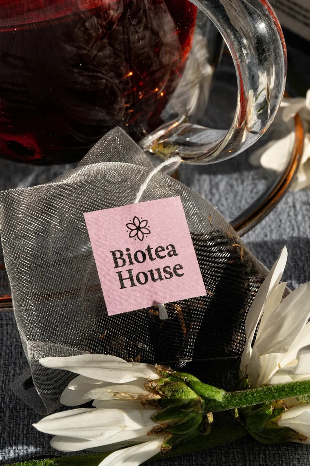 Bolsita piramidal transparente de Biotea House con su etiqueta rosada junto a una copa de vidrio con té rojo y flores blancas, bajo luz cálida.