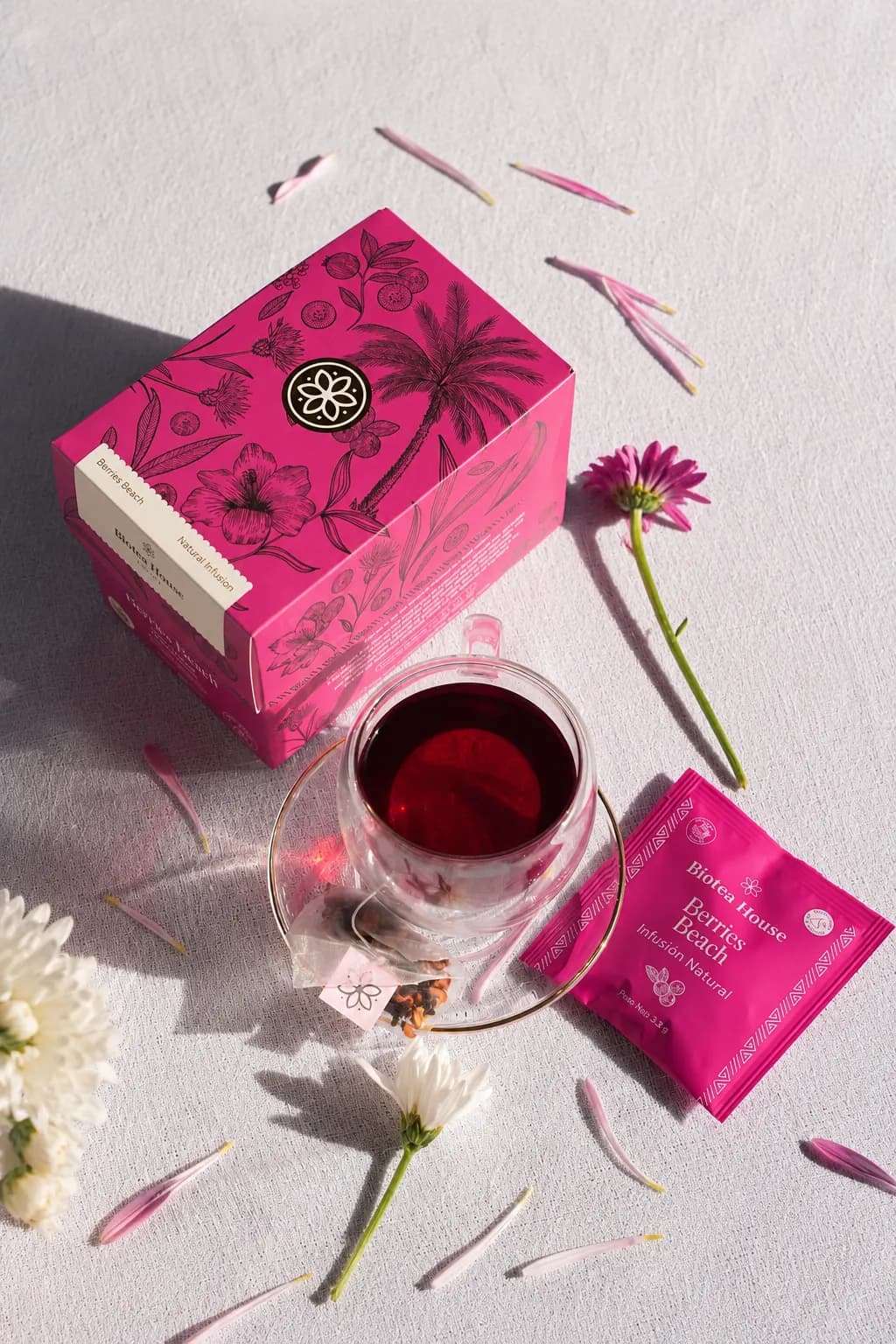 Caja de Berries Beach de Biotea House en tono magenta intenso junto a una taza de vidrio con te rojo y petalos de gerbera esparcidos.